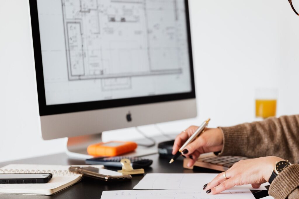 A designer's hands sketching on paper at a desk, with a floor plan open on a computer screen