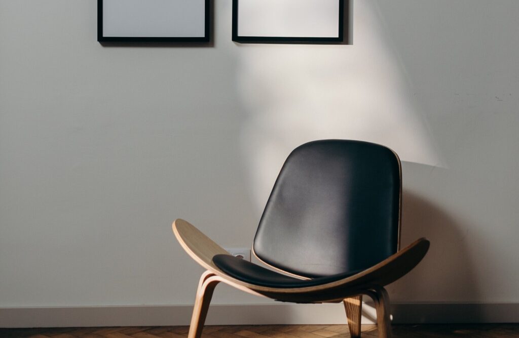 Lounge chair in bent plywood with black leather upholstery against a white wall, under two black picture frames, on a herringbone parquet floor