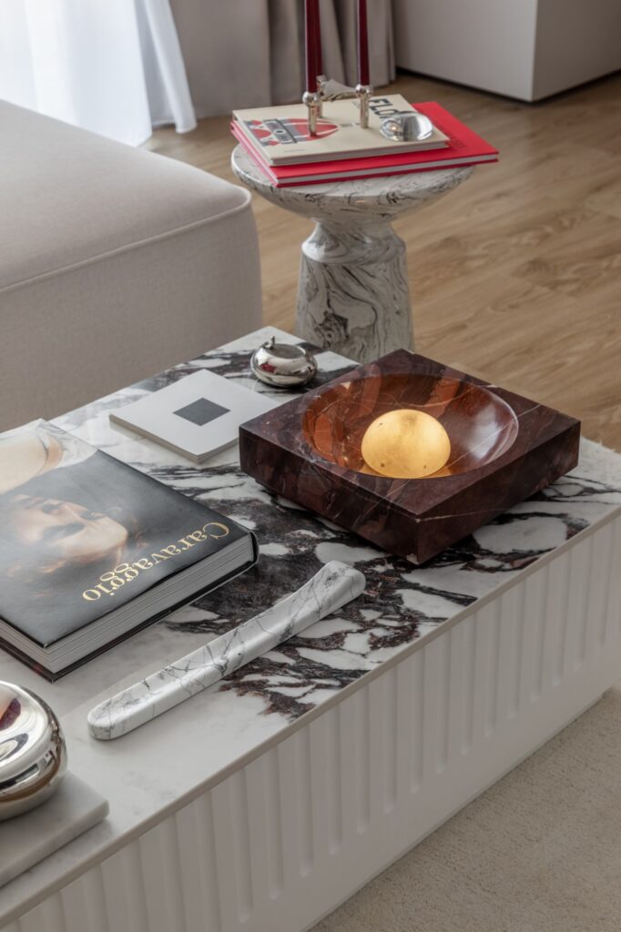 Coffee table with a white and burgundy veined marble top on a fluted white base, with a Caravaggio book, a red marble bowl holding a glowing alabaster lamp and a marbled side table stacked with books behind
