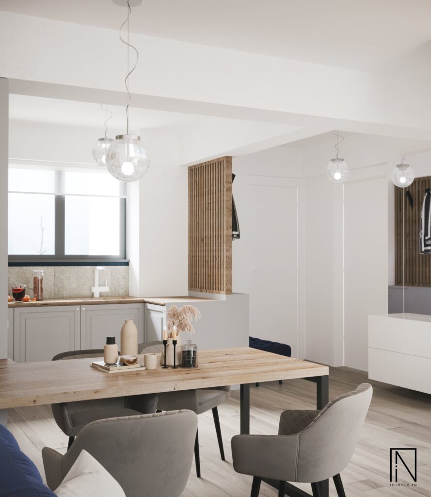 Zona de dining cu masă din lemn rustic pe cadru negru, scaune gri din catifea matlasată și lustre din sticlă, cu bucătăria în spate