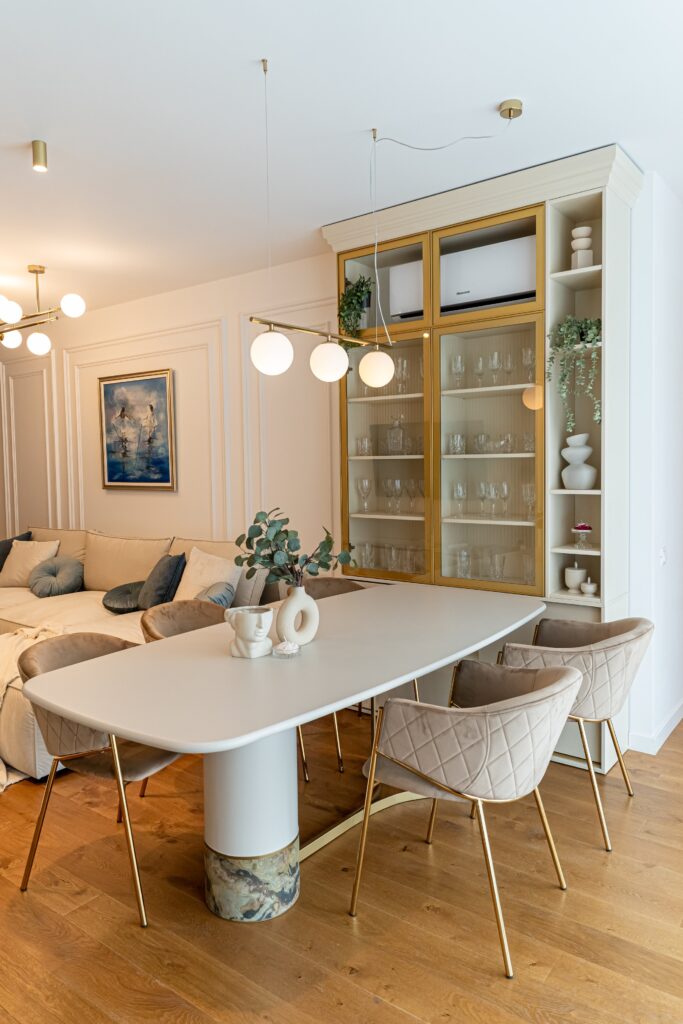 Zonă de dining cu masă ovală albă pe bază placată cu marmură, scaune matlasate gri-bej, pendule cu globuri și vitrina cu ramă aurie